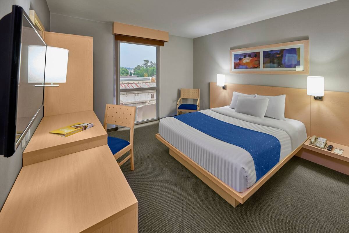 Habitación de hotel con cama, escritorio, silla y ventana con vista a edificios.