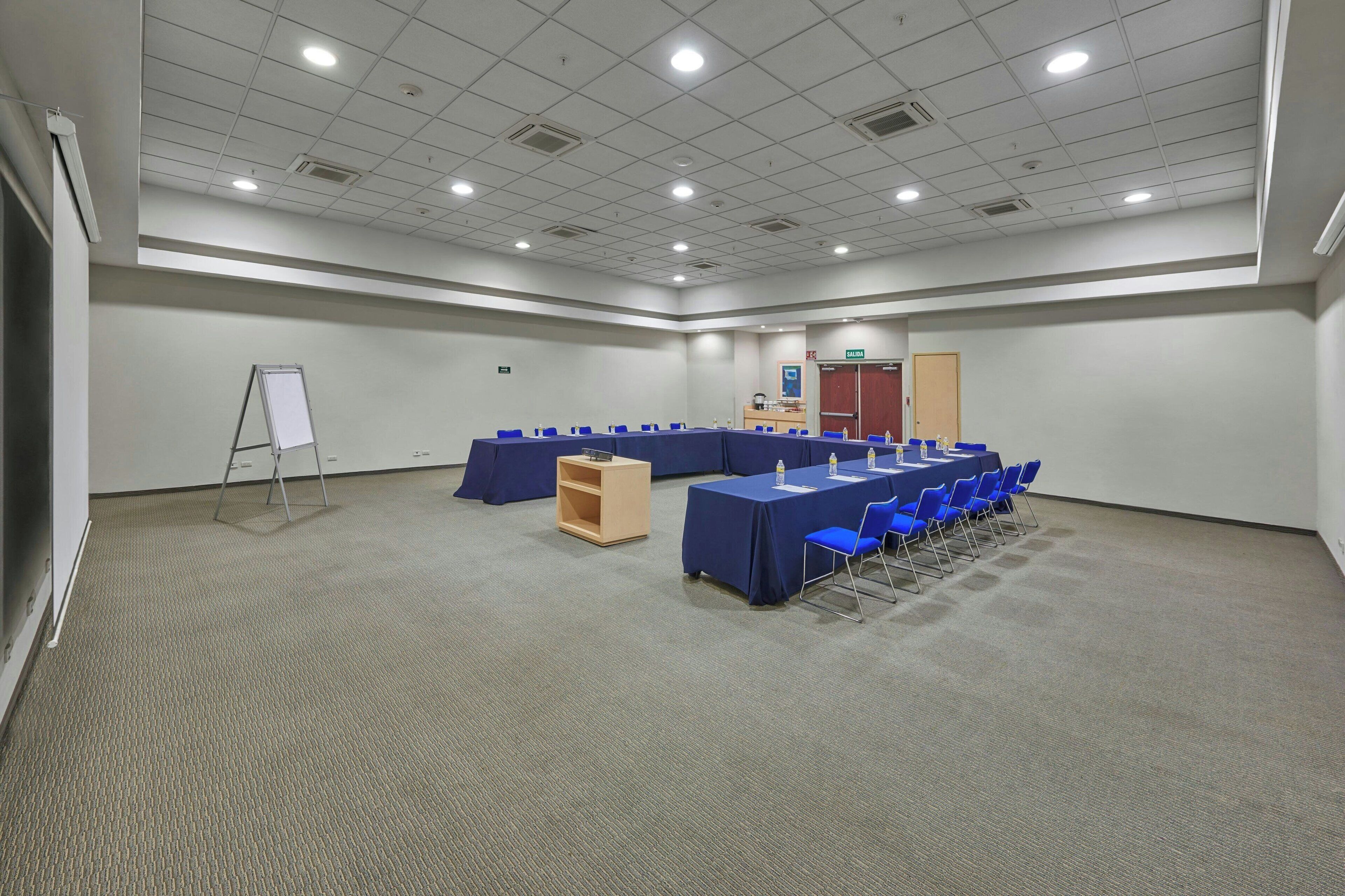 A large conference room with a long table, blue chairs, a whiteboard, and a small wooden cabinet.
