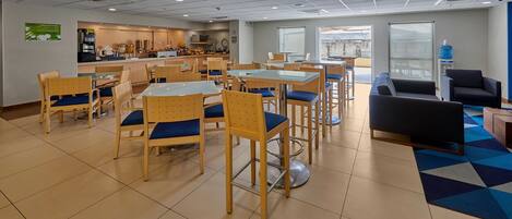 A spacious dining area with multiple tables and chairs, a kitchen in the background, and a large water dispenser.