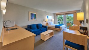 A hotel room with a kitchenette, a blue sofa, a bed with a blue and white bedspread, a wooden desk, and a view of trees through the window. - City Express by Marriott Apizaco (Apizaco)