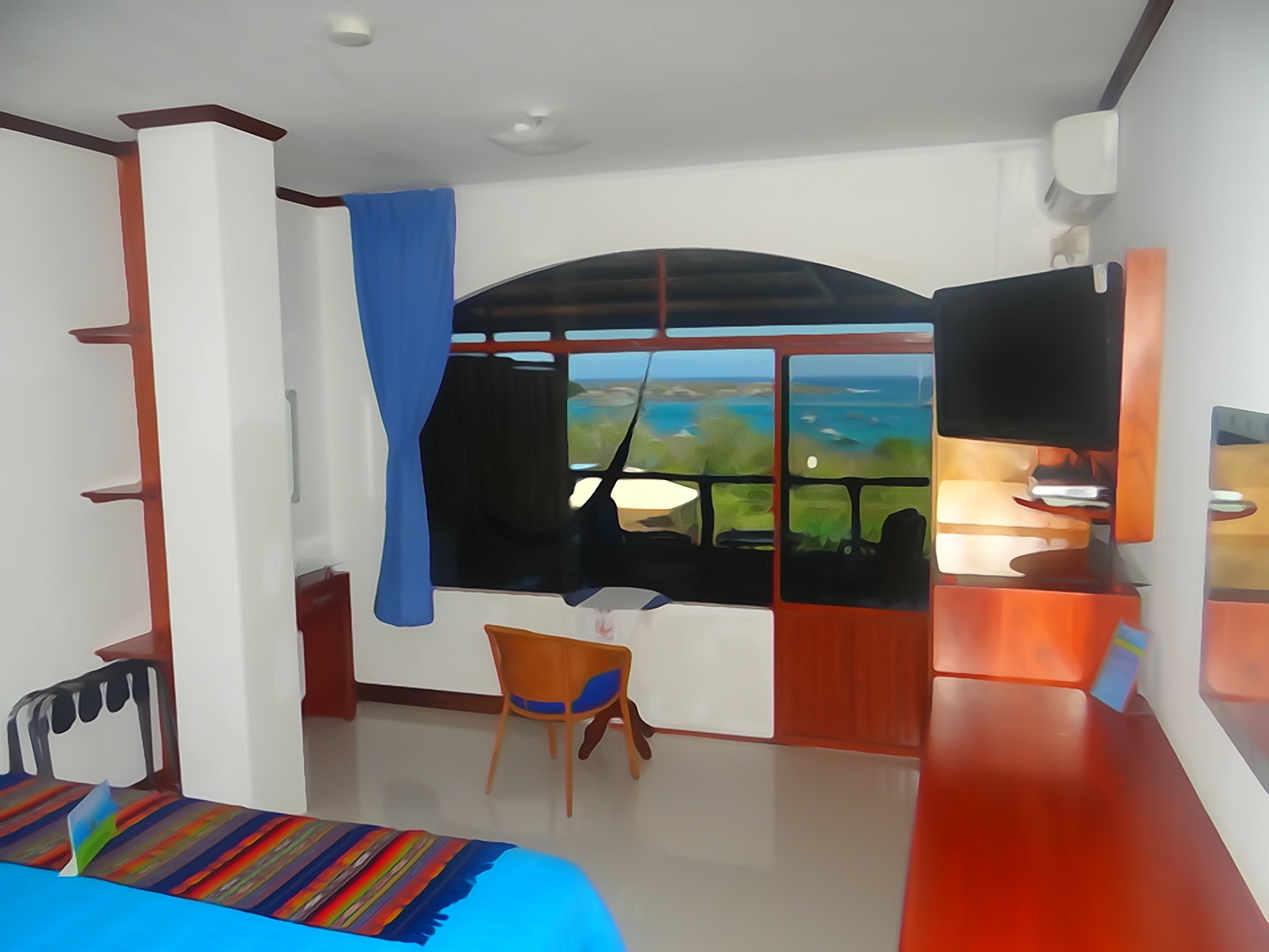 A room with a bed, a TV, a desk, and a window with a view of the sea.