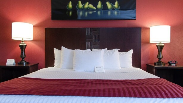 Une chambre d’hôtel avec un grand lit, deux lampes de chevet, un mur rouge et un tableau représentant des poires, accroché au-dessus du lit.