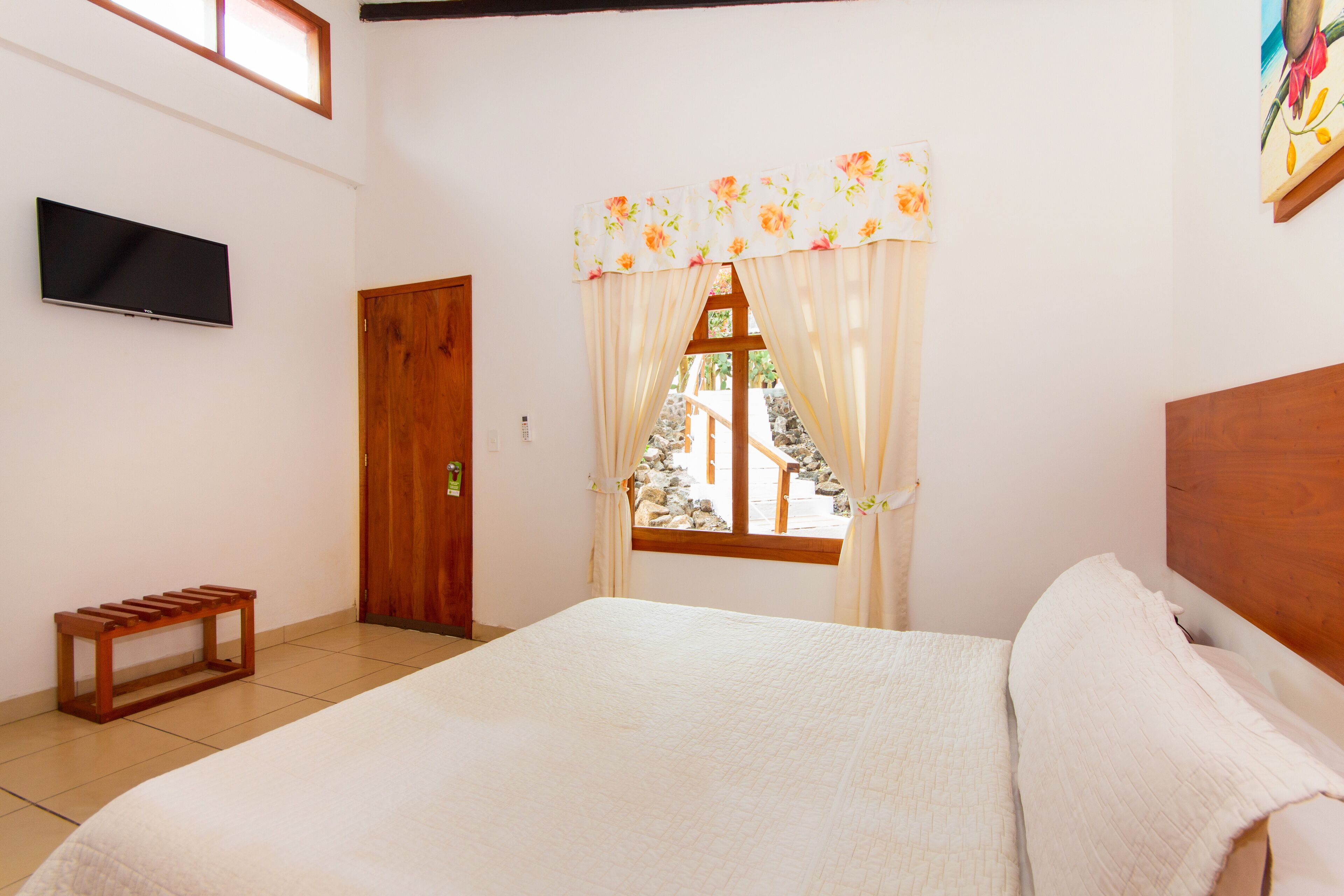 Quarto com cama, televisão na parede, janela com cortinas e um banco de madeira.