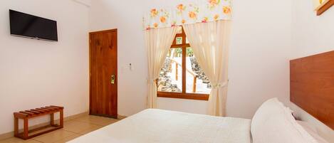 Quarto com cama, televisão na parede, janela com cortinas e um banco de madeira.