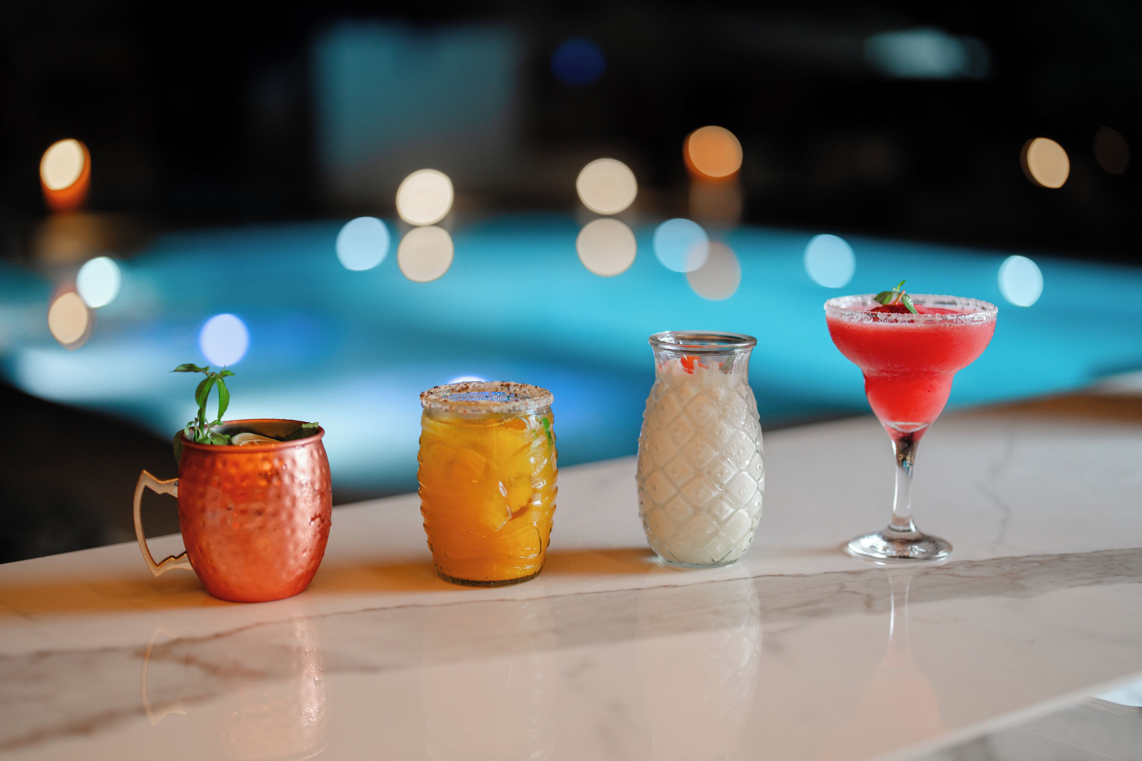 A bar area with four different drinks on a marble surface, a pool with lights in the background.