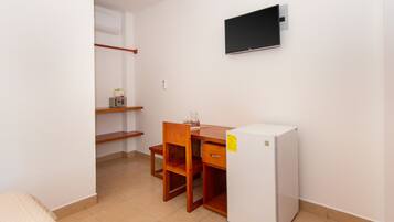 A small, clean room with a wooden desk, a white refrigerator, a TV mounted on the wall, and a shelf with a box and a bottle.
