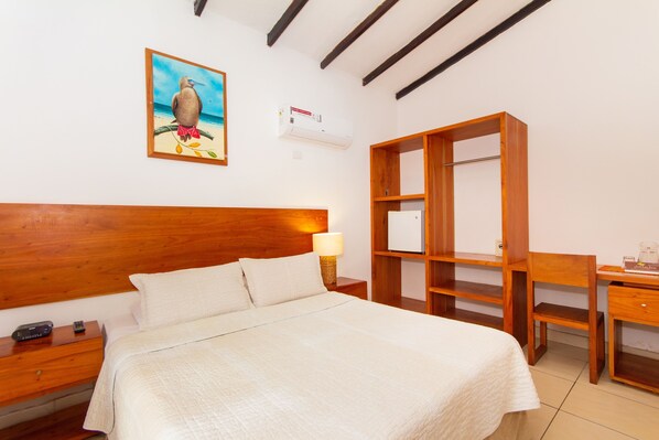 A hotel room with a wooden headboard, a bed with white linens, a nightstand with a lamp, a wooden desk, and a shelf unit. - Hotel Fiesta (Puerto Ayora)