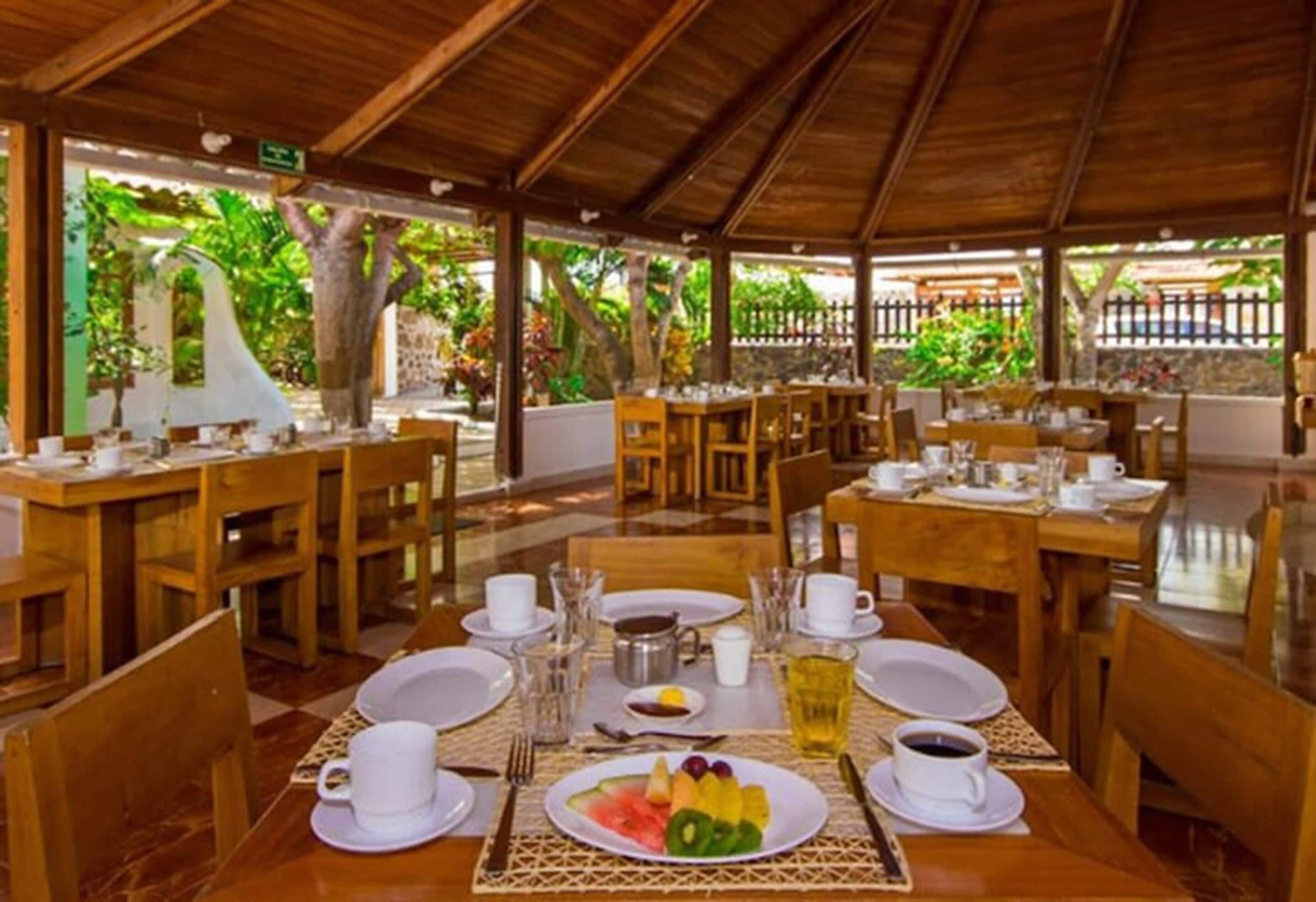 Un espacio exterior cubierto para comer, con mesas y sillas de madera, listo para el desayuno, con platos de fruta y tazas de café.