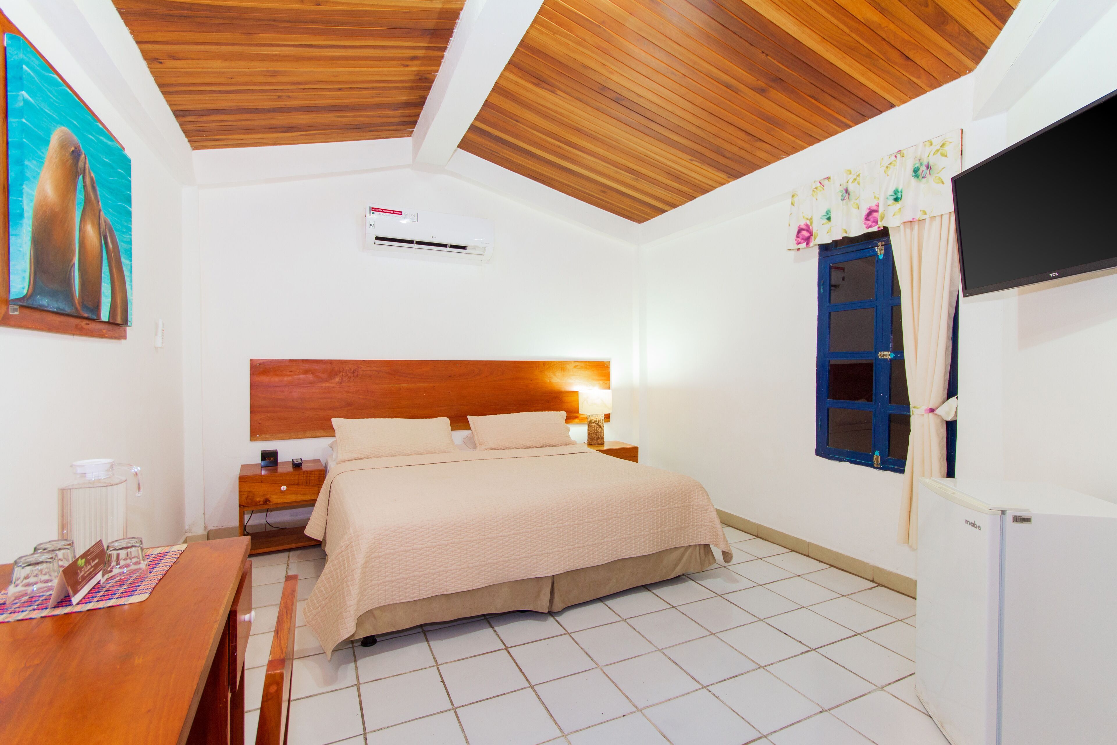 A bedroom with a wooden ceiling, a bed with a beige bedspread, a nightstand with a lamp, a window with curtains, and a wall-mounted TV.