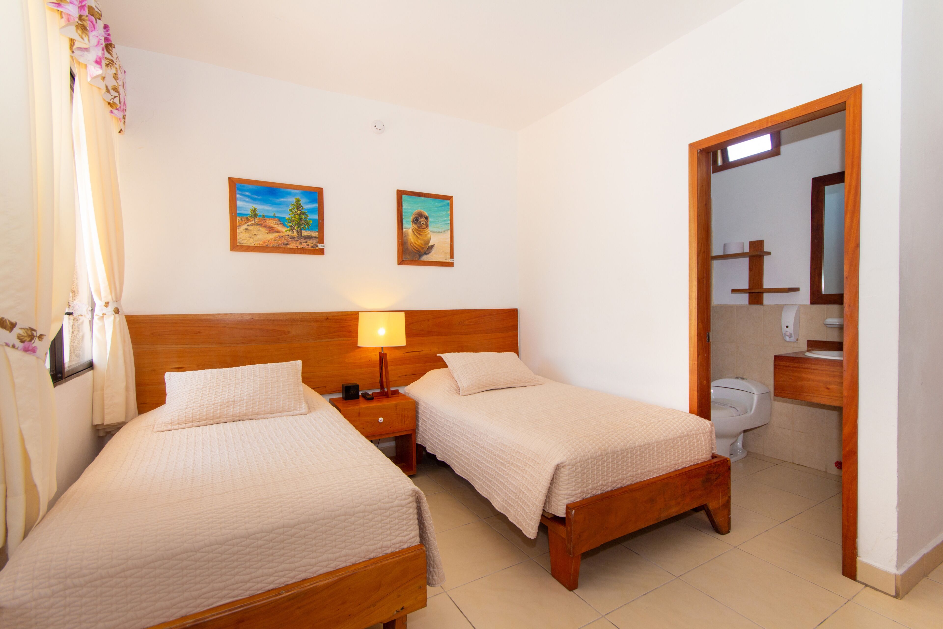 Une chambre avec deux lits, une tête de lit en bois, une table de chevet avec une lampe, et une salle de bain visible à travers une porte ouverte.