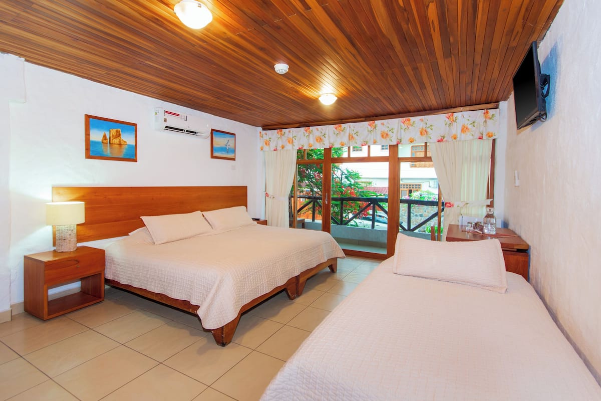 A hotel room with two beds, wooden headboards, a nightstand with a lamp, a TV mounted on the wall, and a balcony with a view of greenery.