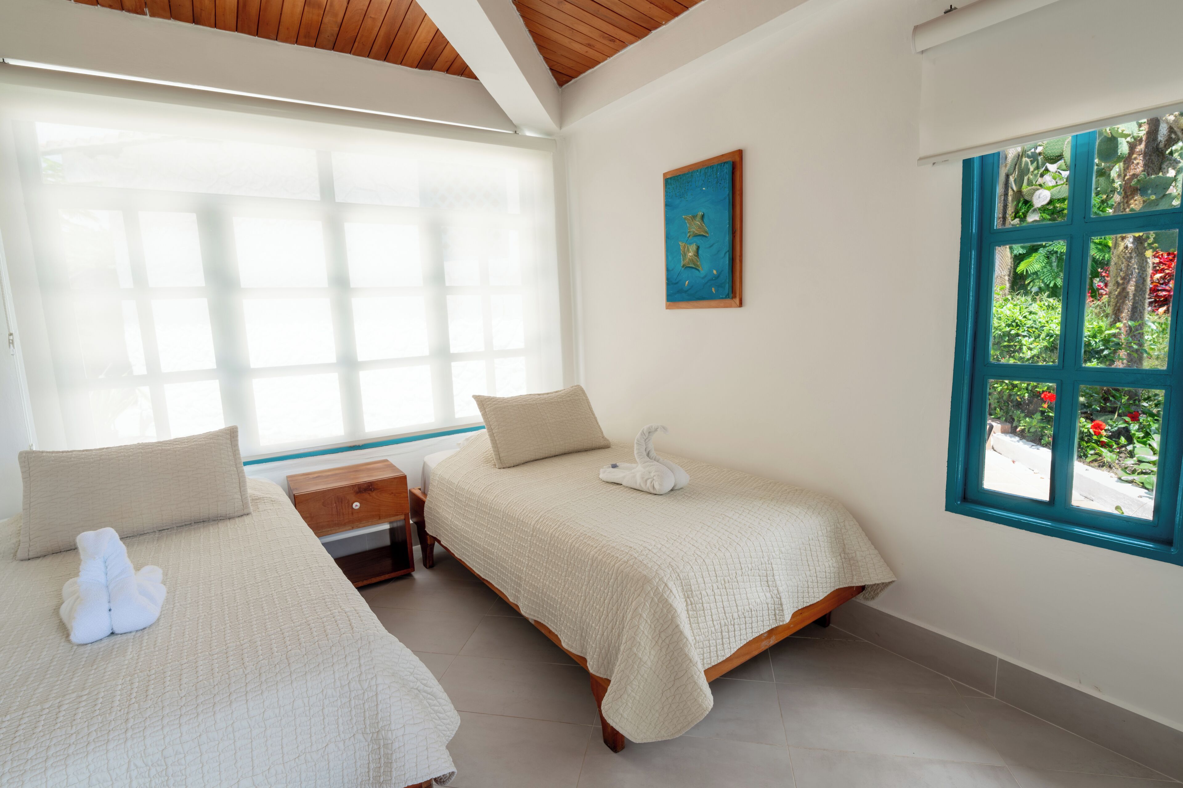 A bedroom with two beds, a wooden nightstand, a window with a view of greenery, and a painting on the wall.
