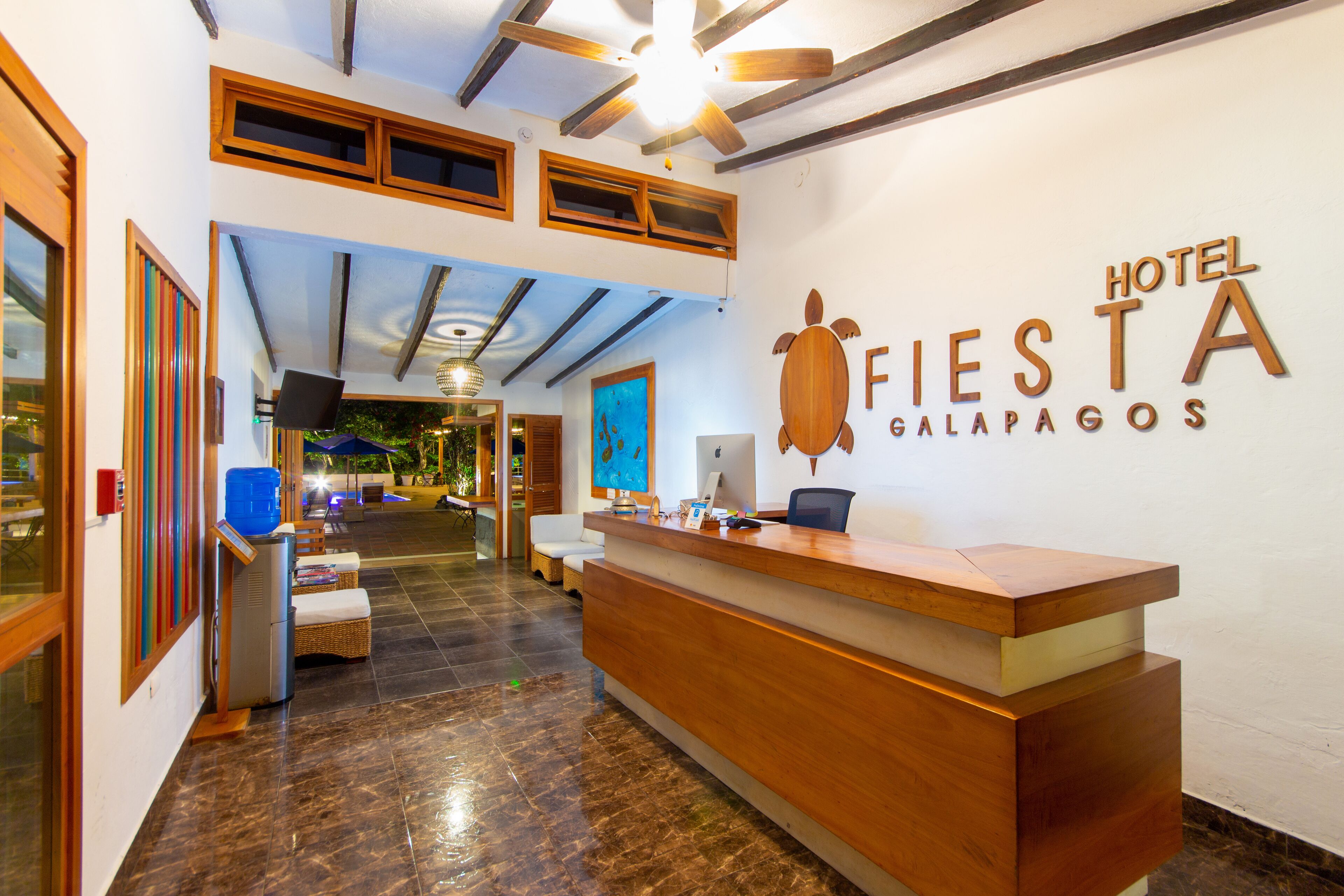 A hotel reception area with a wooden reception desk, a turtle logo, and a sign reading "Hotel Fiesta Galapagos".