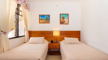 A room with two beds, a wooden headboard, a nightstand with a lamp, and two framed paintings on the wall.