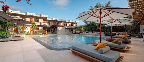 Zona de piscina con sillones, sombrillas y un edificio con balcones.