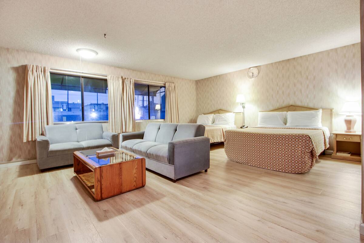 A hotel room with a sofa, a small table, a bed with a patterned bedspread, a nightstand, and a window with curtains.