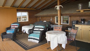 Un chalet confortable avec un intérieur en bois, comprenant un coin repas avec deux tables et des chaises, un coin cuisine avec une machine à café et une grande fenêtre laissant entrer la lumière naturelle.