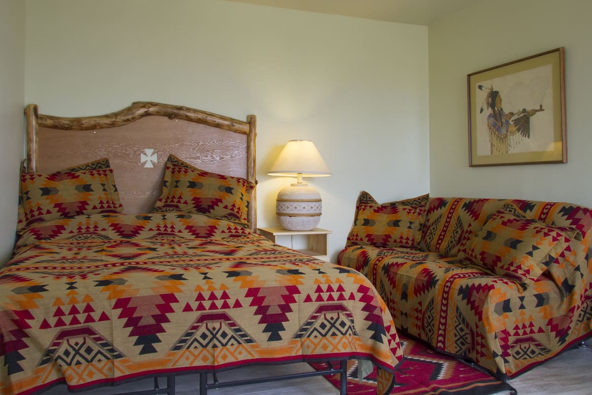A room with a bed and a sofa, both covered with patterned blankets. There is a lamp on a bedside table and a framed picture on the wall.