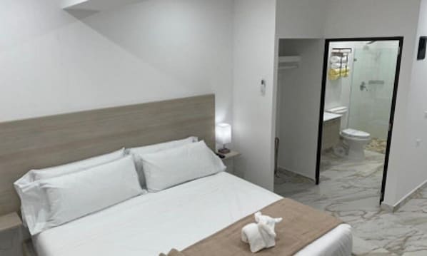 Habitación de hotel con cama, mesita de noche, lámpara y baño visible a través de una puerta abierta.