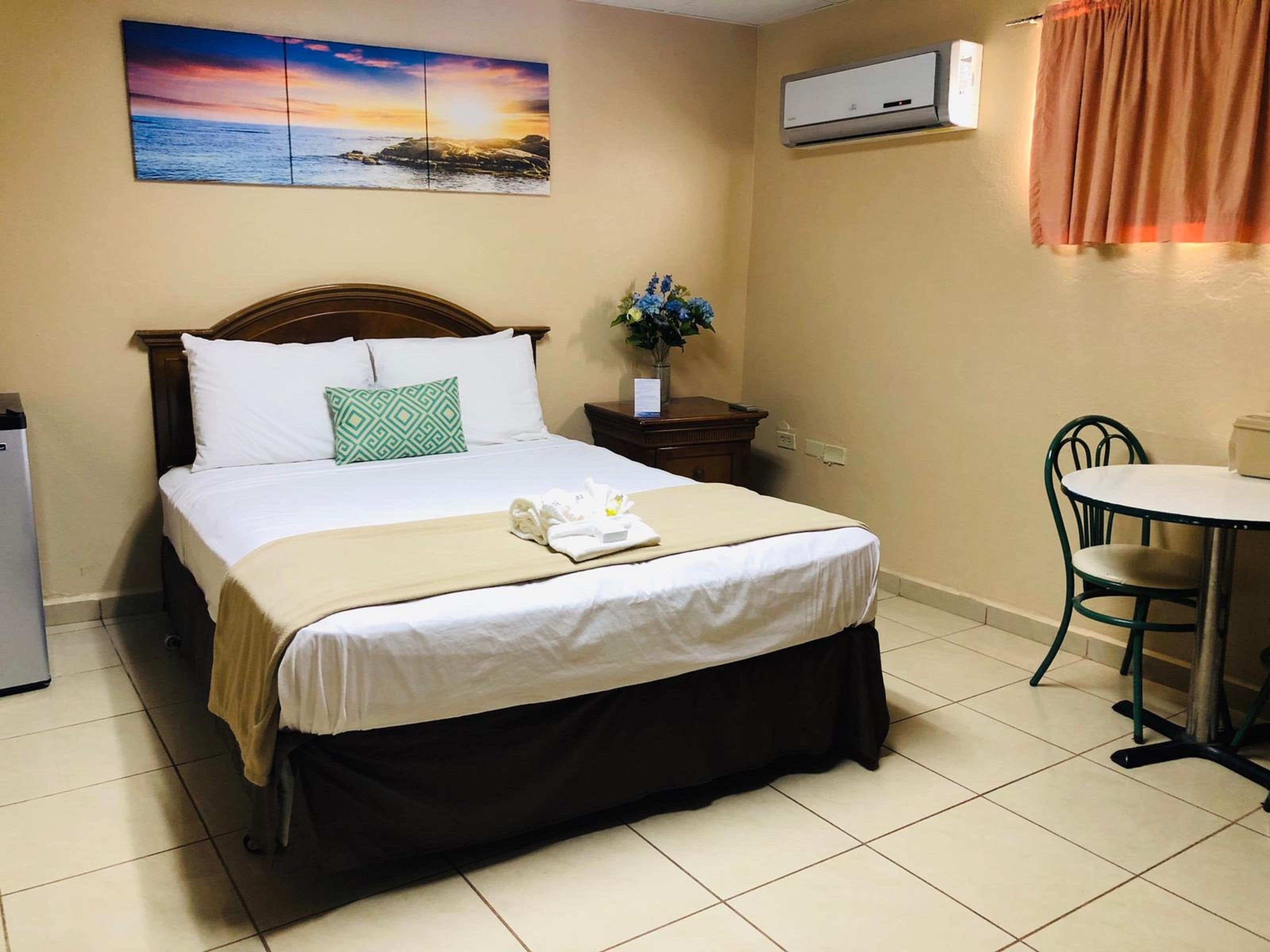 A neatly made bed with a green pillow, a bedside table with a vase of flowers, a small table with a chair, and an air conditioning unit on the wall.