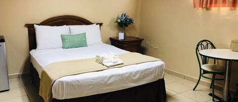Una cama bien hecha con un cojín verde, una mesita de noche con un florero, una pequeña mesa con una silla y un equipo de aire acondicionado en la pared.