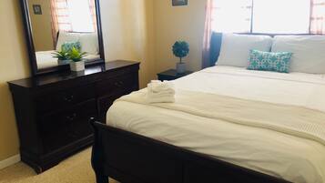 Uma cama bem arrumada com roupa de cama branca, um aparador de madeira escura com gavetas, uma pequena planta em vaso e um espelho sobre o aparador.