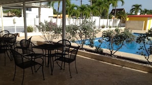 A covered outdoor seating area with tables and chairs, a swimming pool, and a white fence with palm trees in the background. - Arecibo Inn (Arecibo)