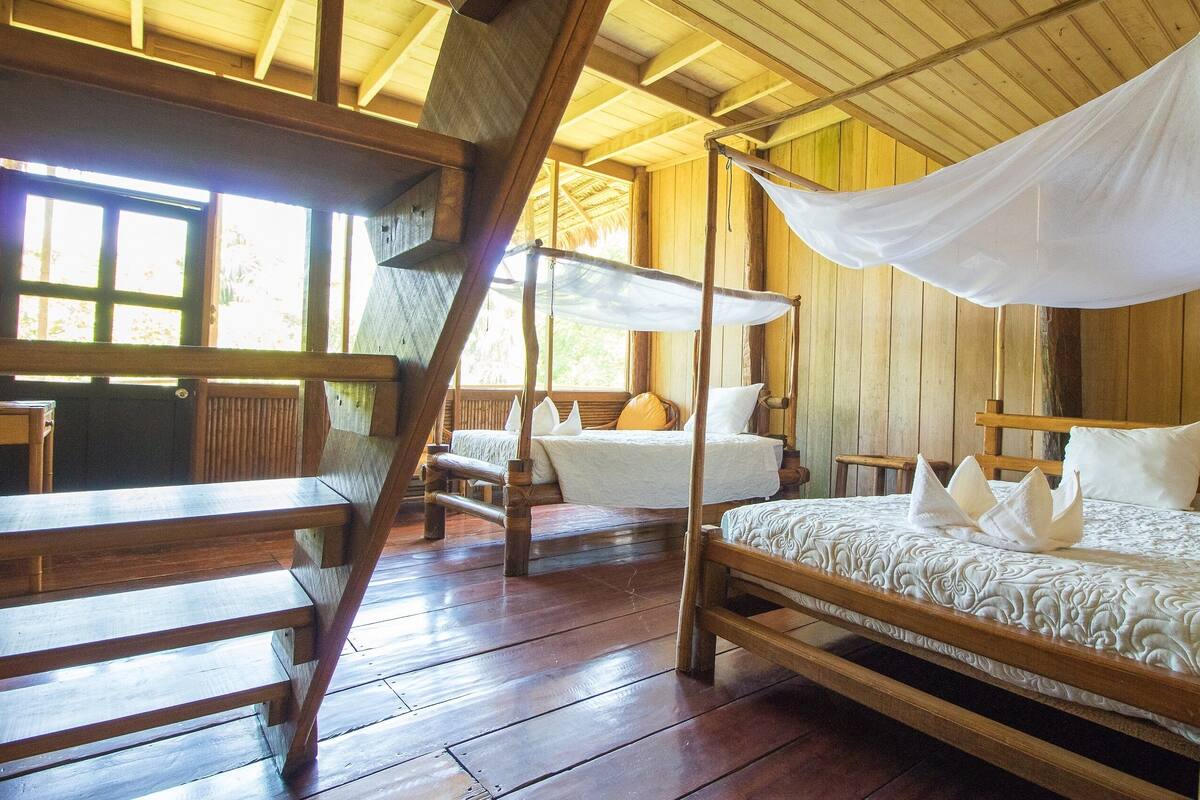 Ein traditioneller Holzraum mit vier Betten, jedes mit Baldachin. Der Raum verfügt über Holzboden und -wände sowie große Fenster für natürliches Licht.