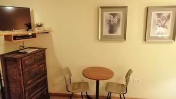 A room with a wooden dresser, a round wooden table with two chairs, a television on a shelf, and two framed pictures on the wall.