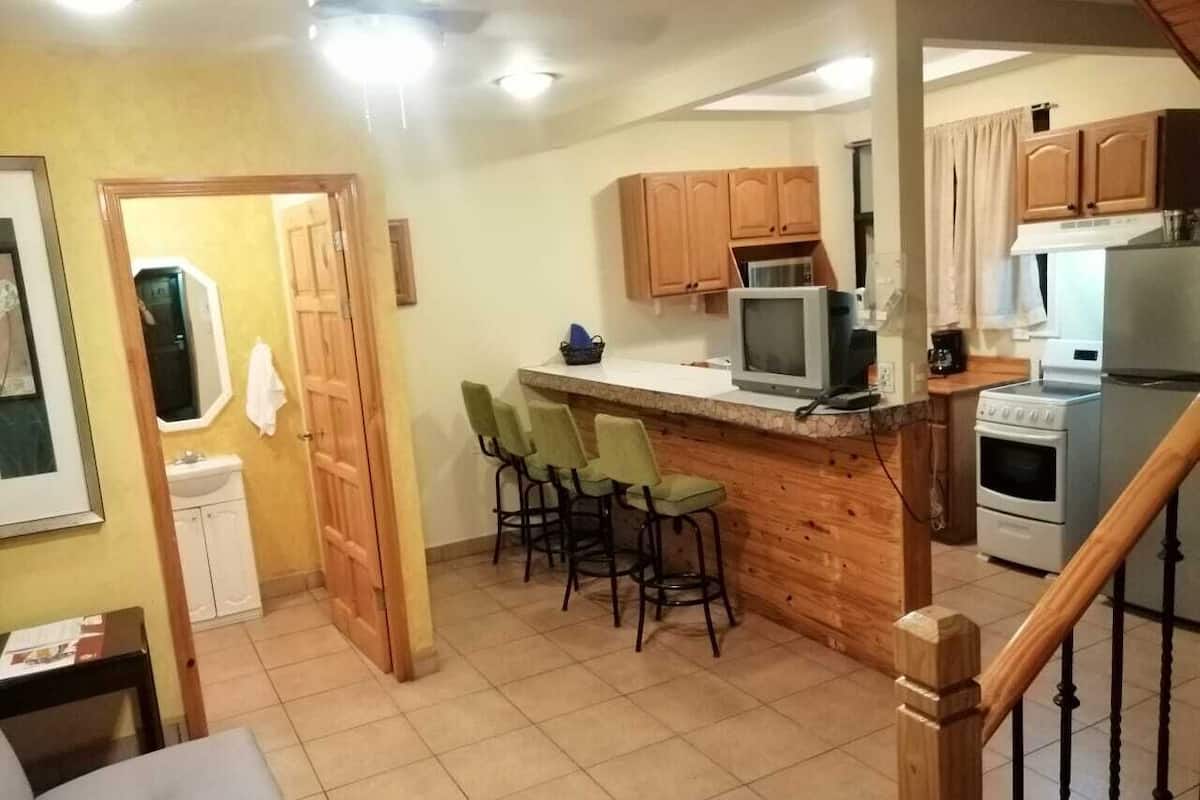 A compact kitchen with wooden cabinets, a built-in microwave, and a stove. There are three bar stools and a small dining table.