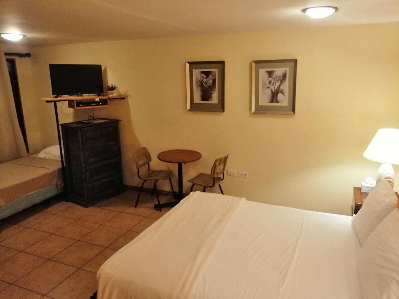 A hotel room with a bed, a TV on a stand, a small table with chairs, and two framed pictures on the wall.