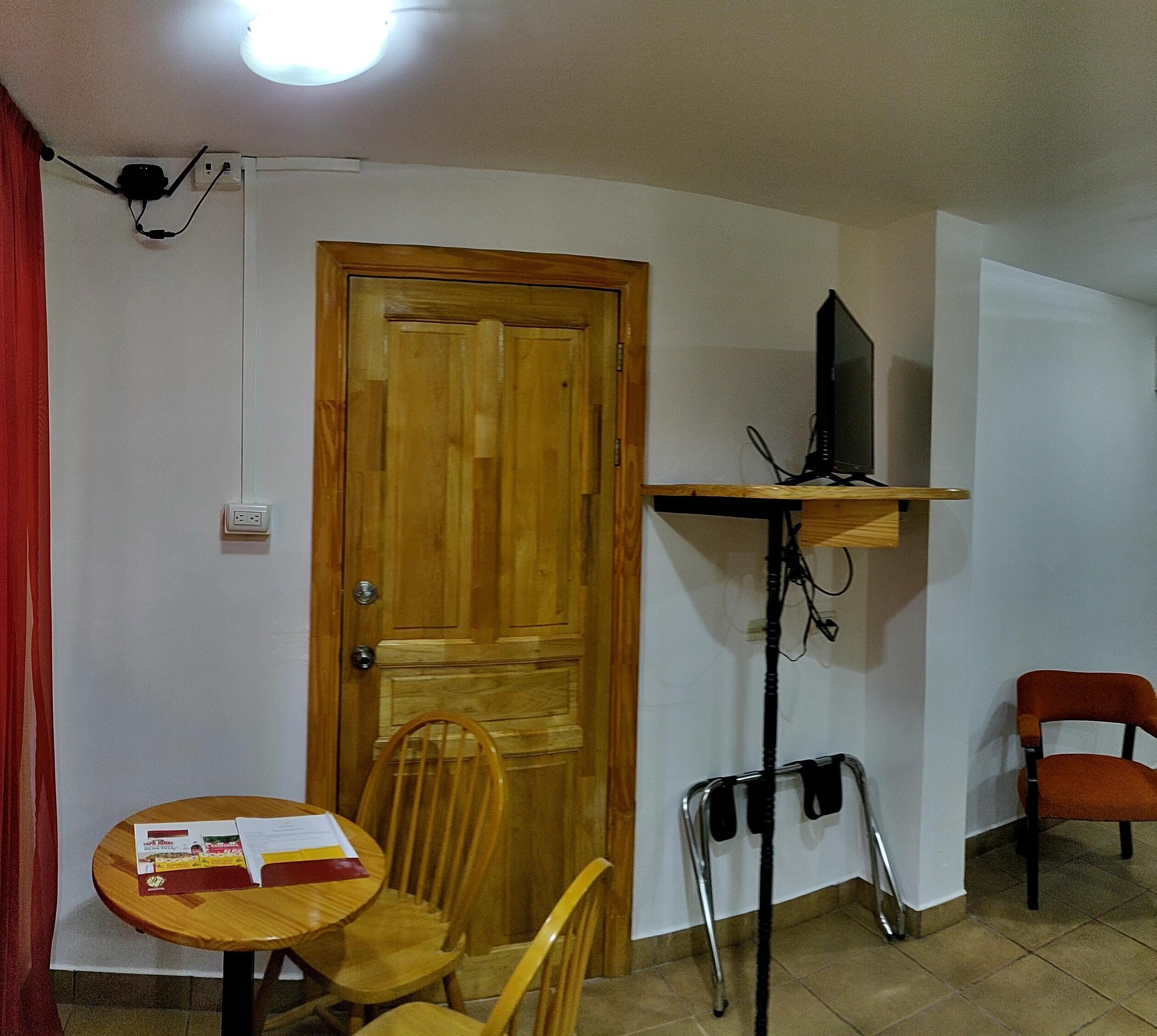 A room with a wooden door, a television on a stand, a table with chairs, and a red curtain.