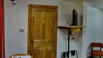 A room with a wooden door, a television on a stand, a table with chairs, and a red curtain.