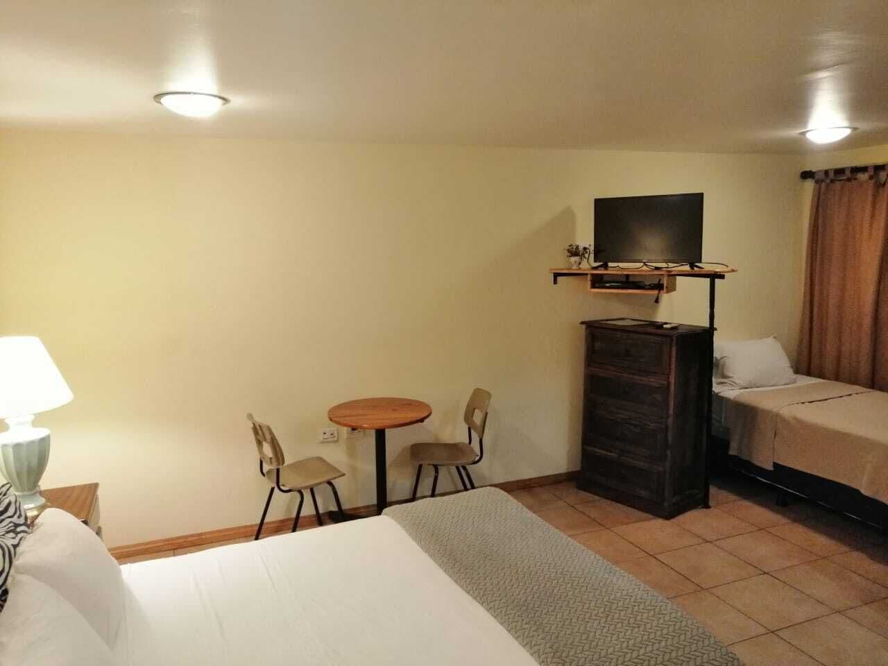 A hotel room with a bed, a desk with a lamp, a TV on a stand, and a small table with chairs.