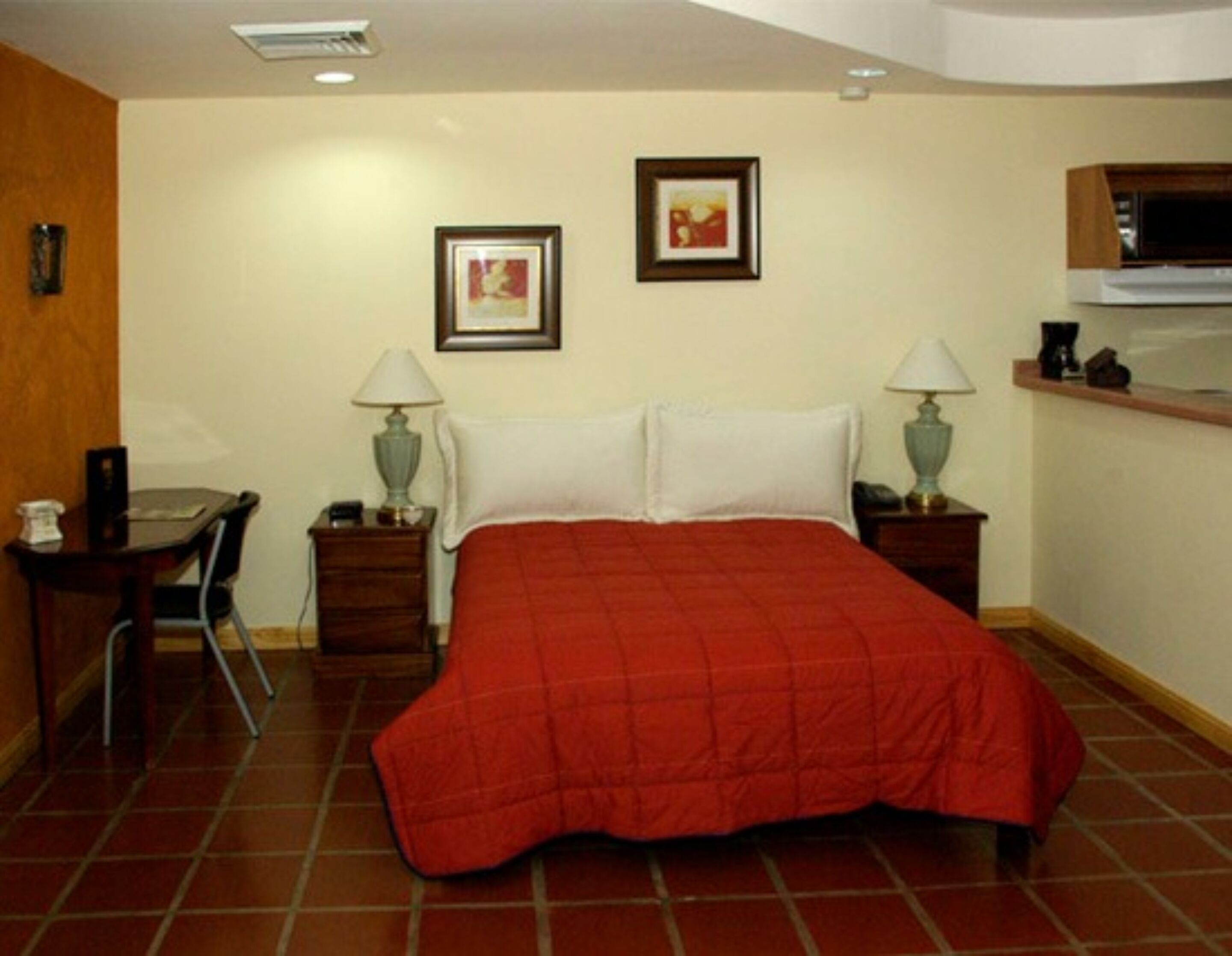 A hotel room with a bed, two bedside tables with lamps, a small desk with a chair, and a kitchenette area.