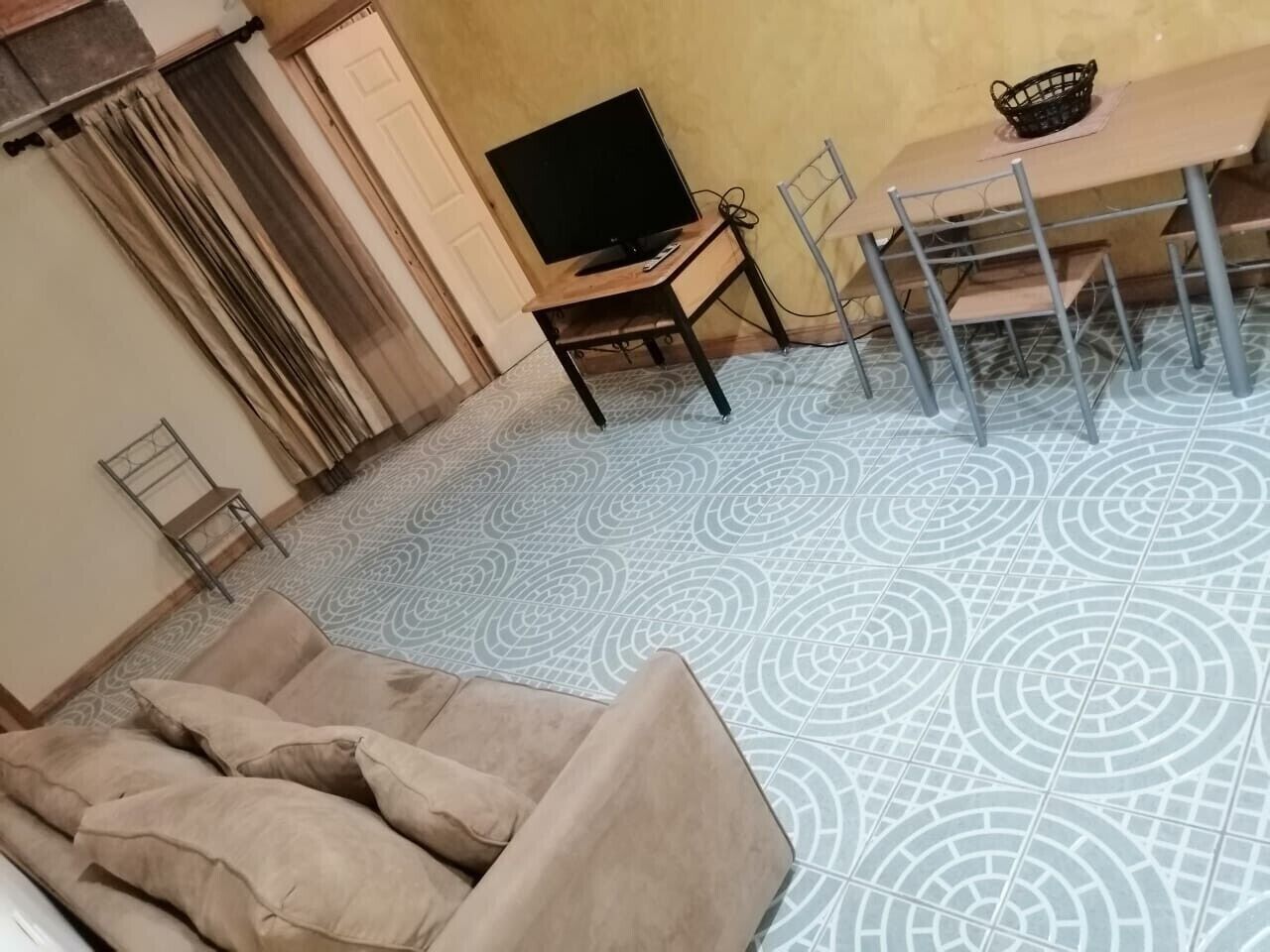 A living room with a sofa, a TV on a wooden stand, a dining table with chairs, and patterned floor tiles.