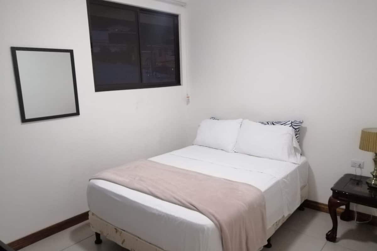 A bedroom with a bed, a window, a bedside table with a lamp, and a framed picture on the wall.