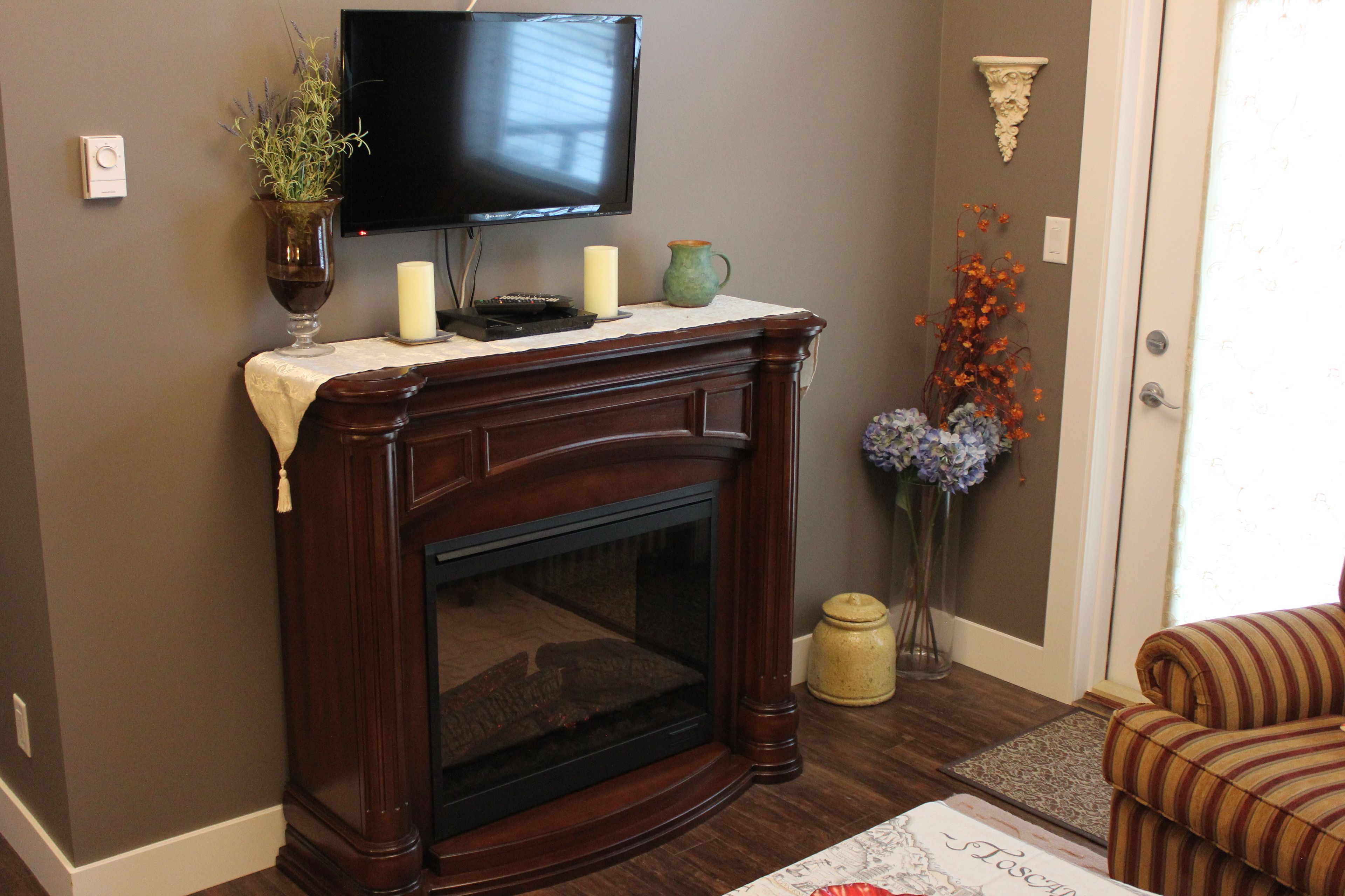 A living room with a dark wood fireplace, a flat-screen TV mounted above it, a vase with flowers, and a striped armchair.