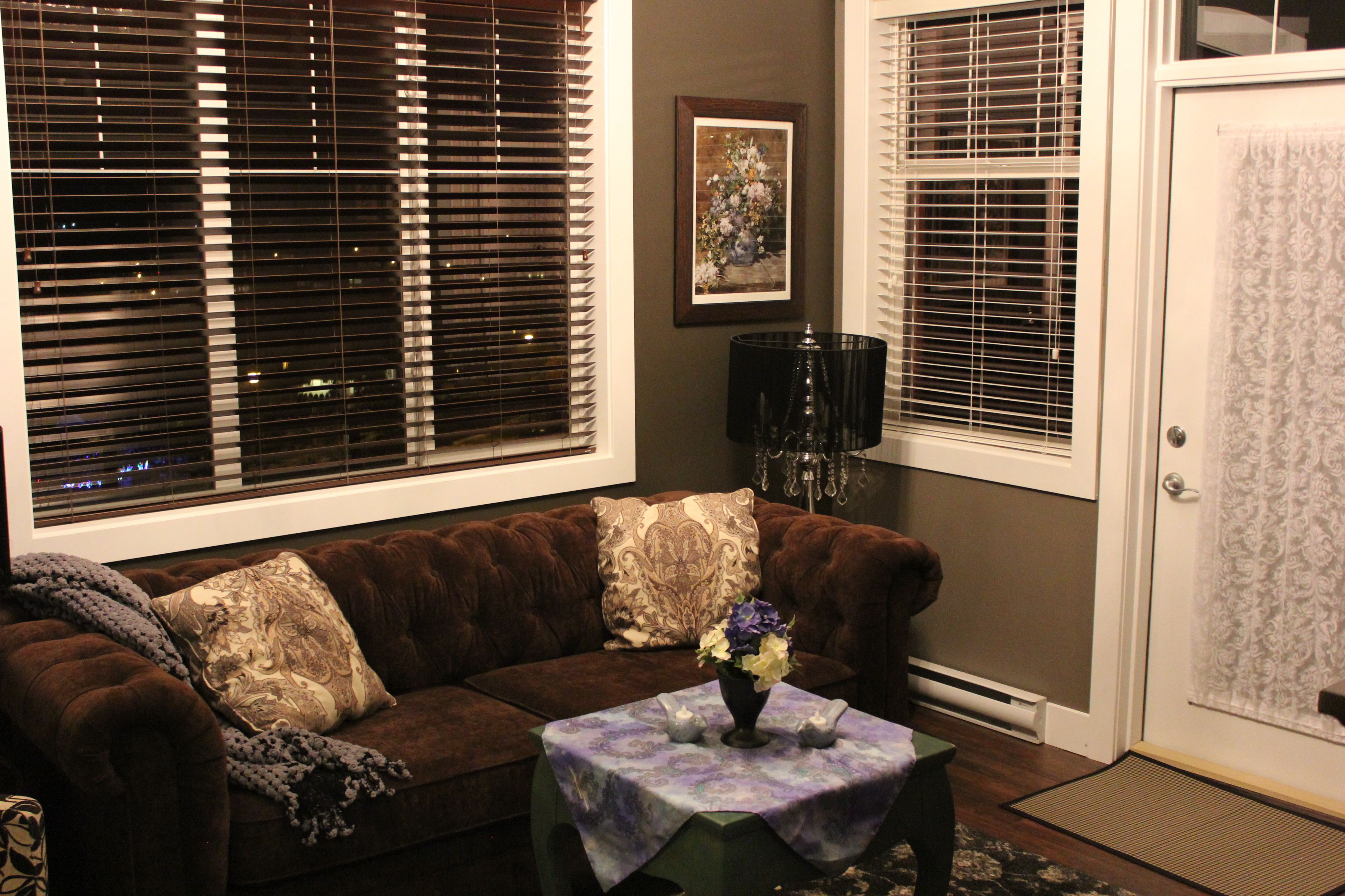 A living room with a brown sofa, two decorative pillows, a small table with a vase of flowers, and a framed picture on the wall.