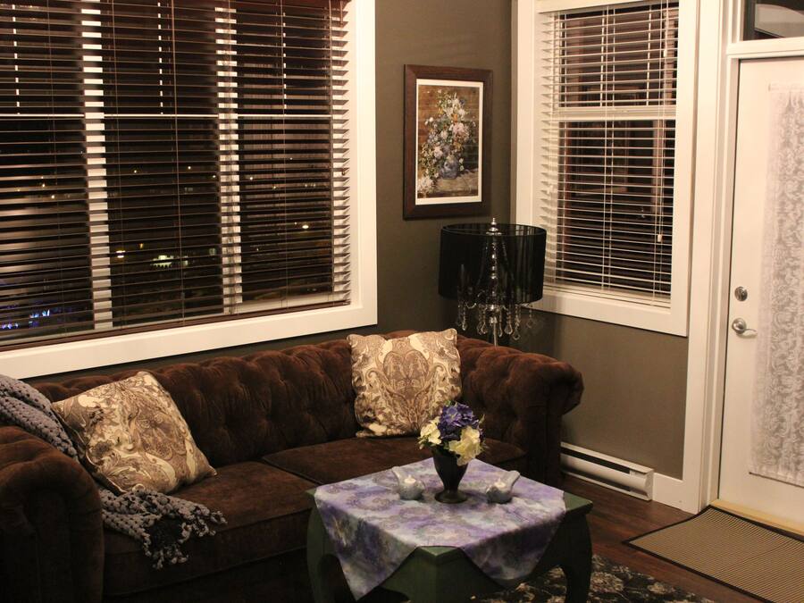 A living room with a brown sofa, two decorative pillows, a small table with a vase of flowers, and a framed picture on the wall.