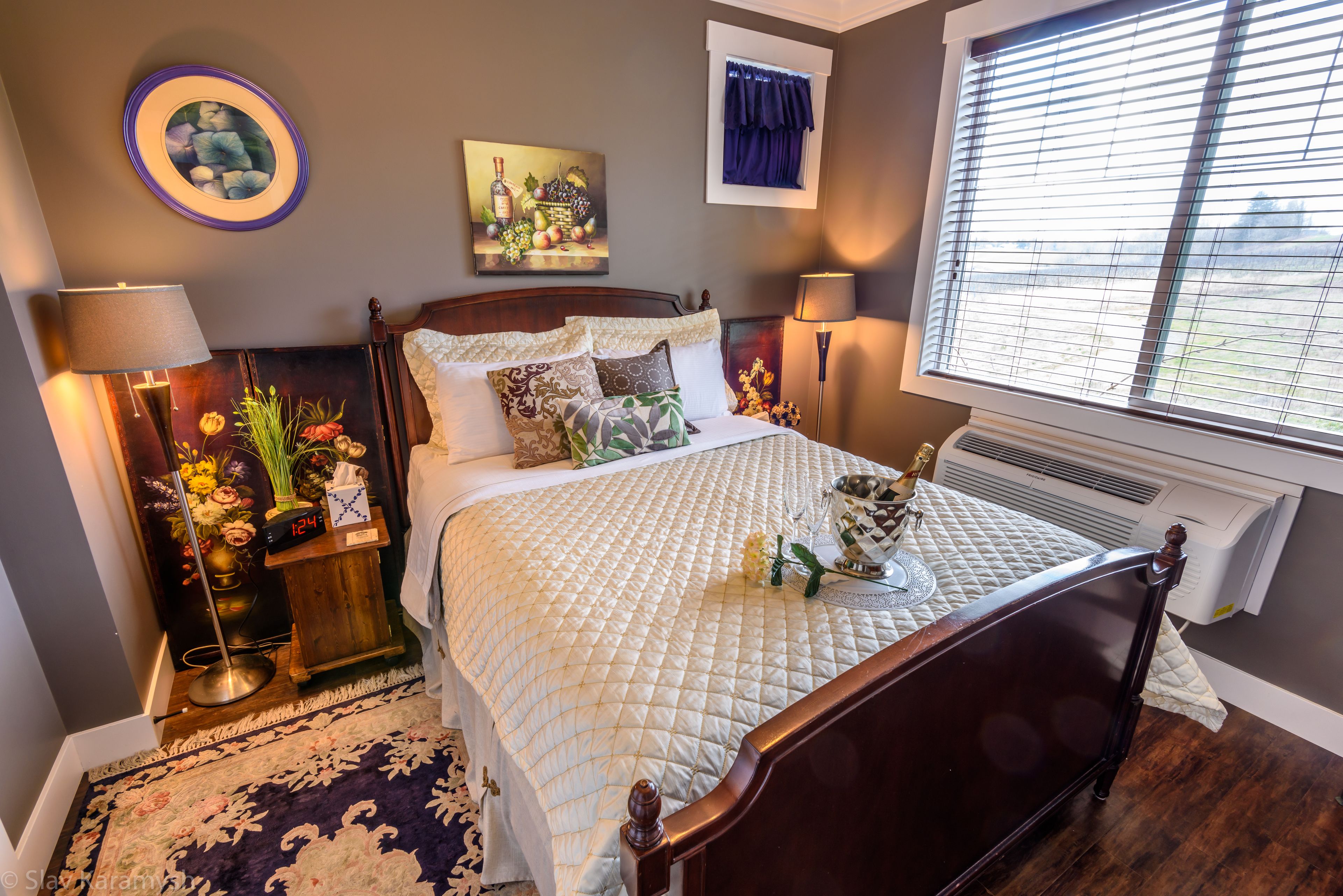 A neatly made bed with a quilt, a bedside table with a lamp, a vase of flowers, and a painting on the wall.