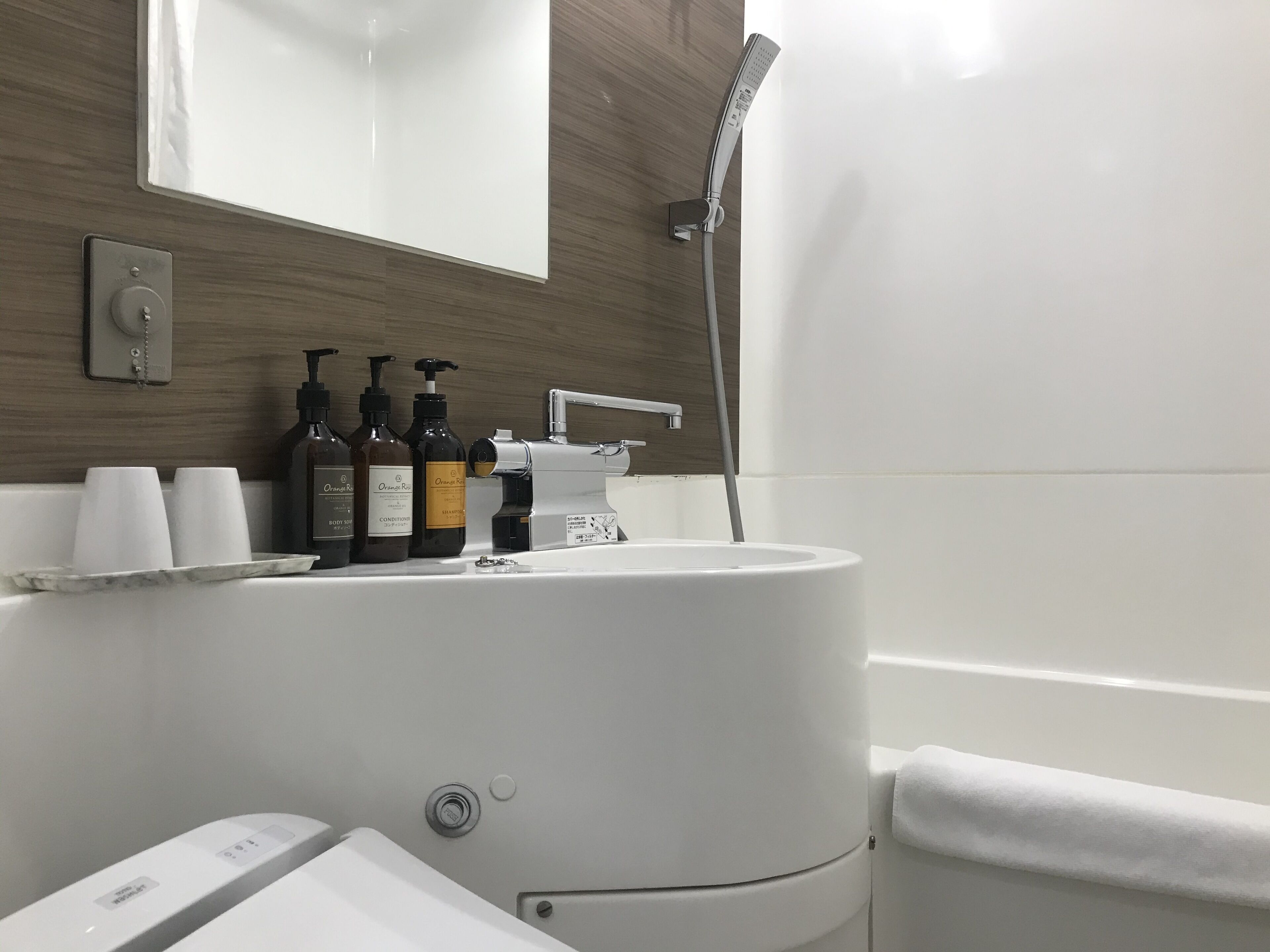 A modern bathroom with a white sink, a mirror, and a shower. There are three bottles of orange body wash and two cups on the counter.