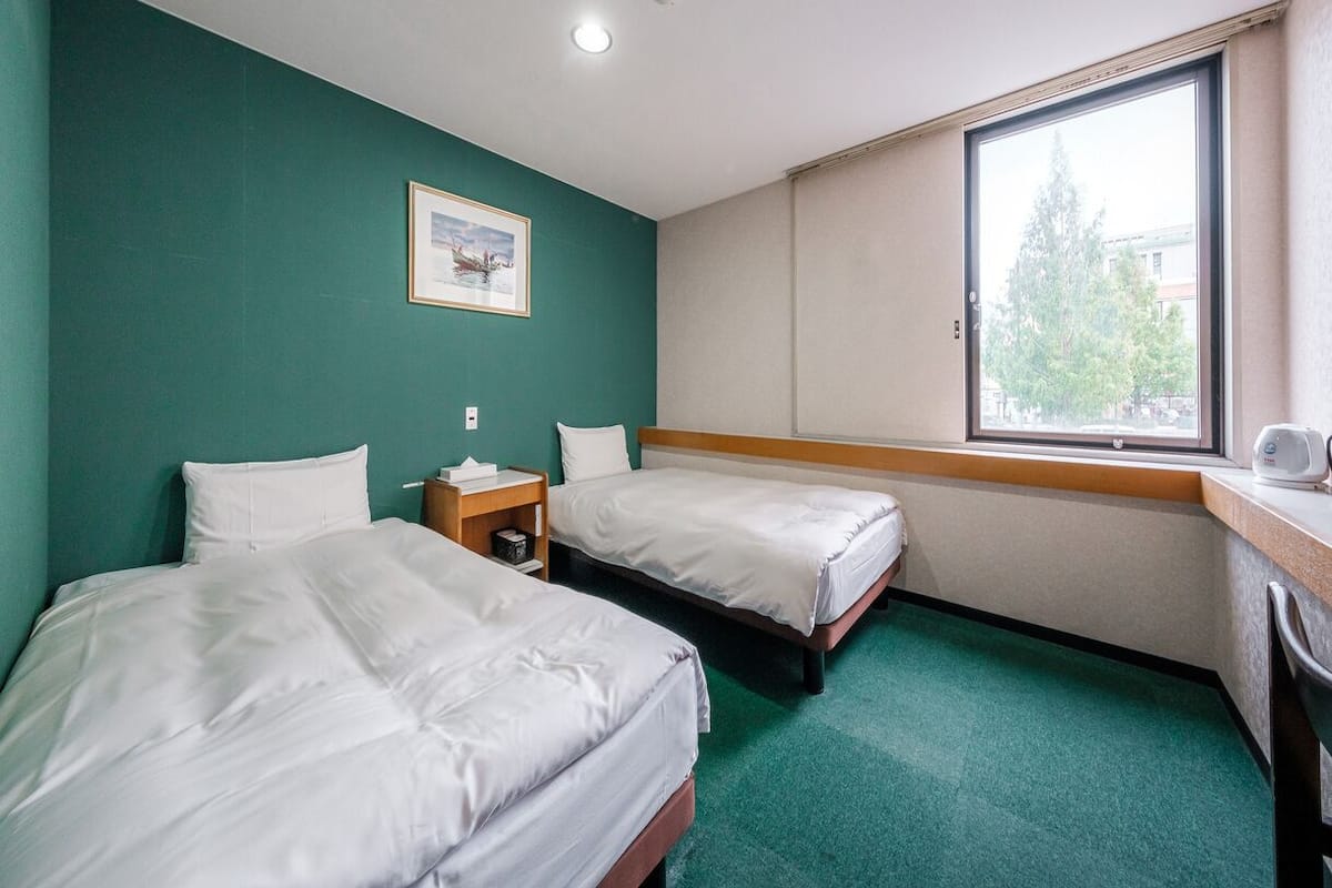 A hotel room with two single beds, a small bedside table with a lamp, a window with a view of trees, and a framed picture on the wall.