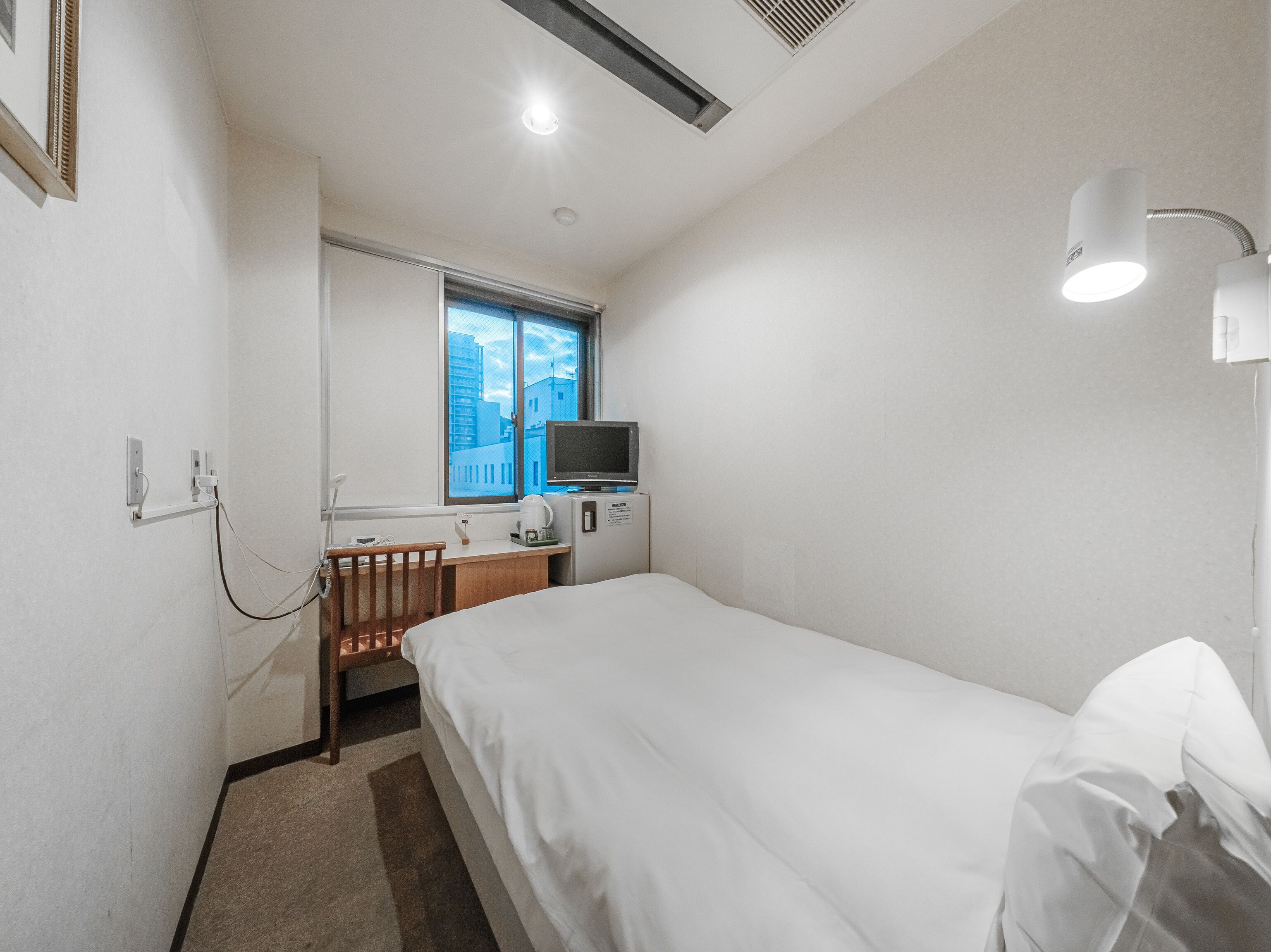Habitación de hotel con cama, escritorio con televisor, ventana con vista a edificios y luz fijada en la pared.