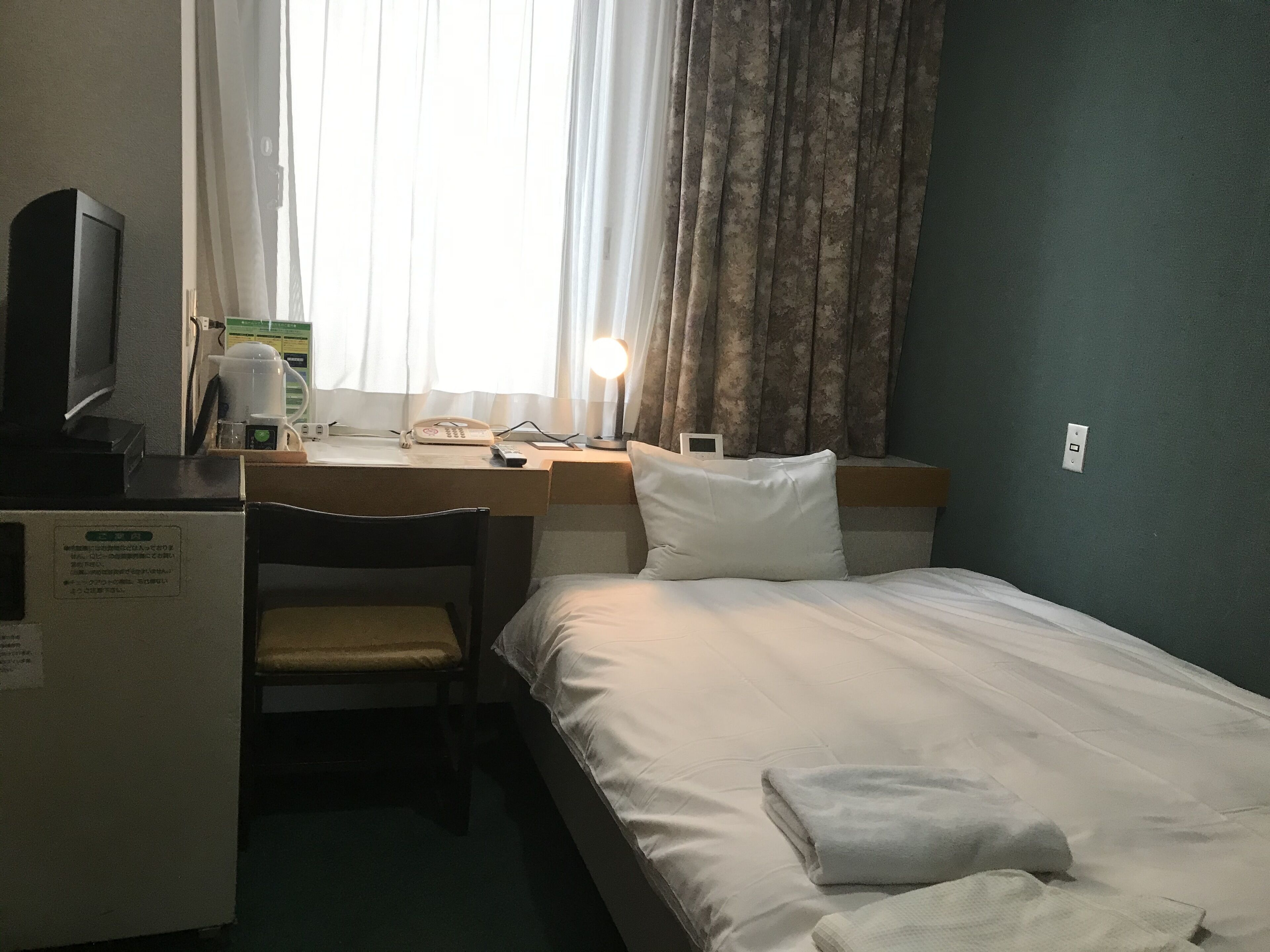 A hotel room with a bed, a desk with a chair, a television, a lamp, and a window with curtains.