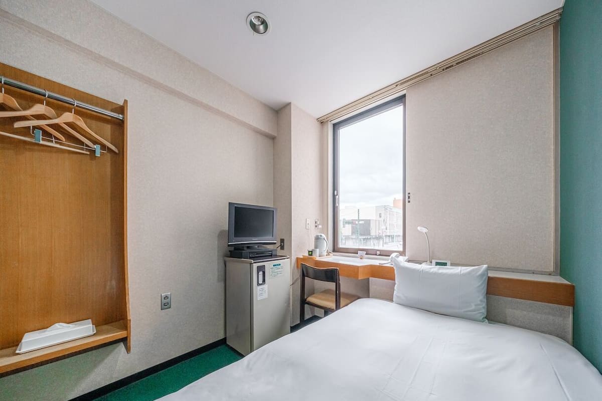 A hotel room with a bed, a desk with a chair, a television on a small cabinet, a wardrobe, and a window with a city view.