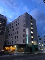Un hotel de varios pisos con una fachada moderna, grandes ventanales y una entrada destacada con un letrero iluminado en verde.