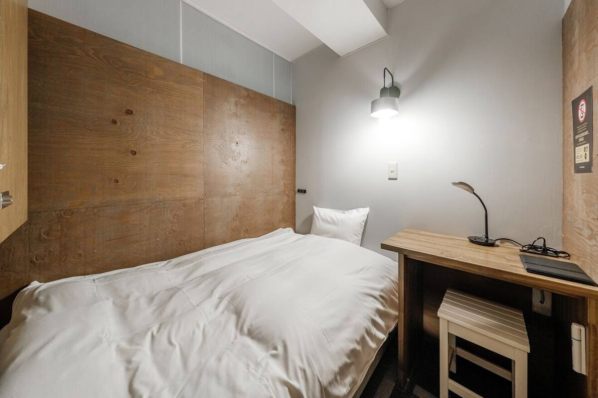 A bedroom with a wooden headboard, a bed with white bedding, a desk with a lamp, and a small bench.