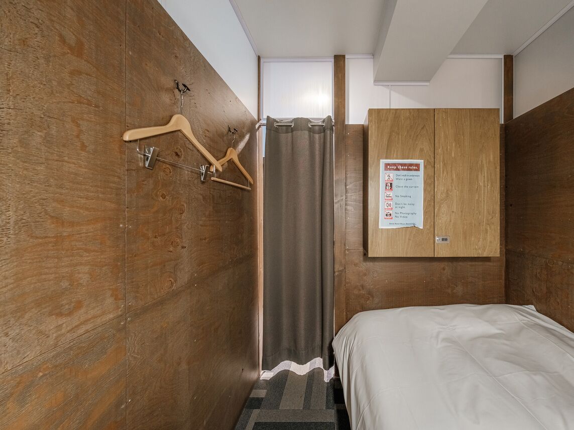 A small, wooden-finished room with a bed, a shower curtain, and a wooden wardrobe. A sign on the wardrobe lists room rules.