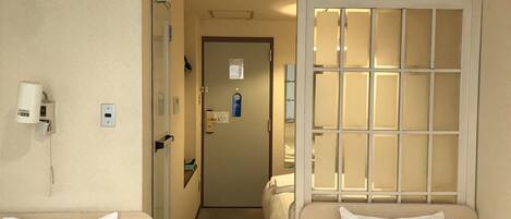Una habitación de hotel con cama, una mesita, una silla, una mesita de noche, una ventana con vistas y una puerta.